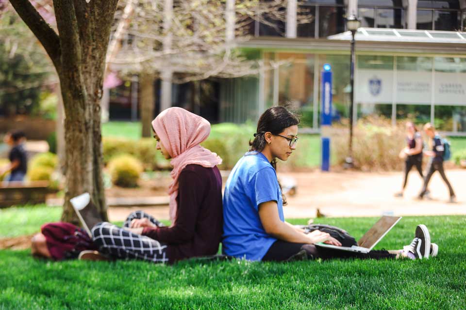 两个学生，一个戴着头巾，背靠背坐在树下的草地上
