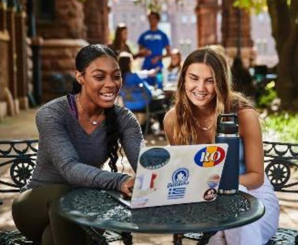 两个面带微笑的学生坐在户外的一张小咖啡桌旁学习，前面放着笔记本电脑